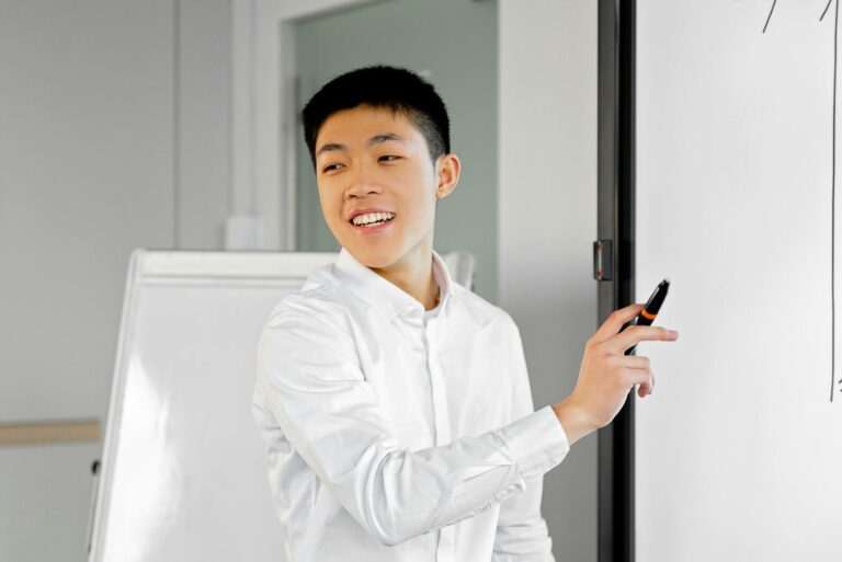 Smiling young man in smart casual attire giving a presentation using a whiteboard indoors.